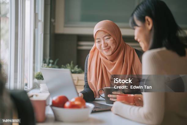 Asian Malay Woman In Hijab With Vitiligo Skin Condition At Kitchen Using Laptop Talking To Her Female Friend Stock Photo - Download Image Now