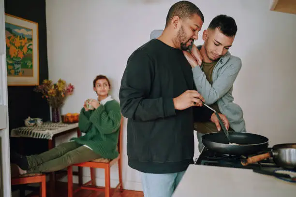 Happy gay couple embracing while cooking breakfast in a kitchen at home with flatmate in the background. LGBTQIA Lifestyle
