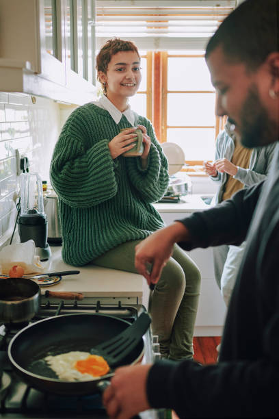 Non-binary person sitting on a kitchen counter talking to flatmate cooking breakfast at home Non-binary person drinking tea and in discussion with flatmate while preparing food in a kitchen at home flatmate stock pictures, royalty-free photos & images