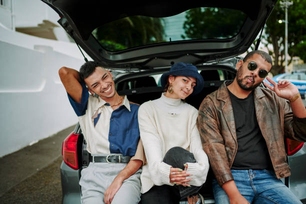 Portrait of friends with a cool attitude making faces and posing looking at camera Happy LGBTQIA friends making facial expressions and posing while sitting on a parked car in the street just before a road trip queer man facing camera stock pictures, royalty-free photos & images