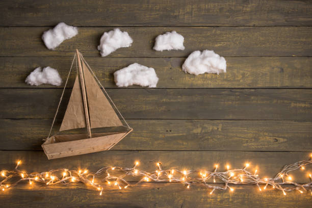 Travel and adventure creative concept - toy boat on a wooden background with cotton clouds. Christmas lights as a sea waves stock photo