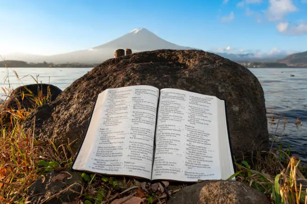 Holy Bible open to Psalms 121, 122, 123 and 124 leaning against a rock on the shore of Lake Kawaguchi against the backdrop of the majestic Mount Fuji, Japan.