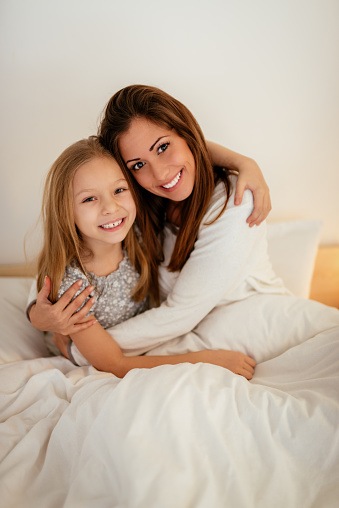 Beautiful smiling mother and her daughter hugging in bed. They are sitting on bed at pajamas and looking at camera.