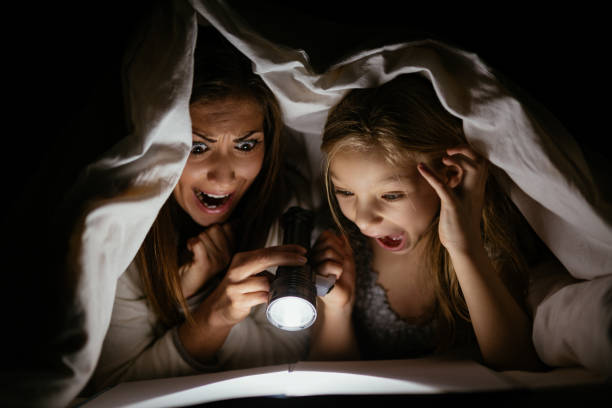 Mother And Daughter Reading Scaring mother and her daughter reading book under bed cover and holding a flashlight. cute horror stock pictures, royalty-free photos & images