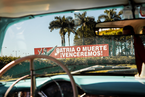 View of a roadside billboard related to the Cuban revolution. The picture was taken in a moving taxi whilst travelling North of Havana.  The billboard displays a popular slogan of the Cuban revolution (patria o muerte) as well as an image of Fidel Castro.