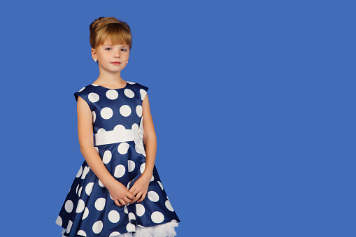 Pretty little girl 8-9 year old in polka dot dress at isolated blue background, looking at camera. Portrait of chic child in image style, isolate on blue. Fashionable kids concept. Copy ad text space