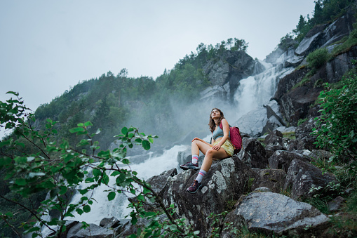 Young Caucasian woman  sitting near the powerful  waterfall in mountains during her hike in Norway