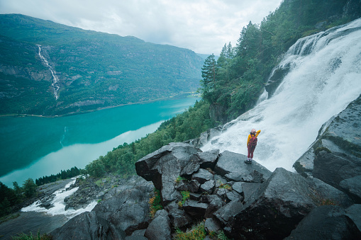 Young Caucasian woman  walking near the powerful  waterfall in mountains during her hike in Norway