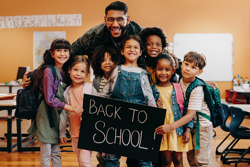 Teacher and his primary school class celebrating coming back to school. Male educator and a group of children smile at the camera while standing in a classroom. School class feeling excited to start a new year.