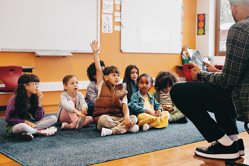 Boy raises his hand to answer a question in a classroom; he is sitting on the floor with other kids and the teacher is sitting in front of the class. Early child development in an elementary school.