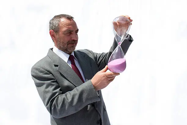 Middle-aged man in a grey suit watching the sand pour through an hourglass.