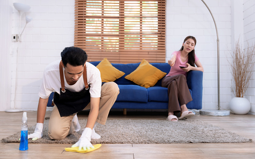 Asian man cleaning floor while lazy girlfriend doesn't help with housework.
