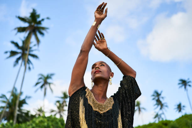 Outrageous black lgbtq person in luxury gown poses with hands up on tropical palm tree location. Non-binary ethnic fashion model in posh outfit stands gracefully at picturesque location. Pride month. stock photo