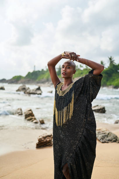 istock Epatage lgbtq black male posing with hands up on camera on scenic ocean beach. Non-binary ethnic fashion model in long posh dress wears jewellery stands gracefully on sea shore and lighthouse. 1489457732