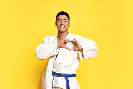 Portrait with happy, cheerful man, taekwondo, karate athlete in kimono showing heart sign amorous feelings over yellow background. Concept of sport, education, skills, martial arts, healthy lifestyle