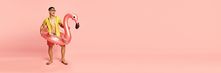 Banner with handsome young student, man wearing beach clothes standing with inflatable flamingo over pink background. Copy space for ad. Concept of human emotions, celebrating, vacation, party