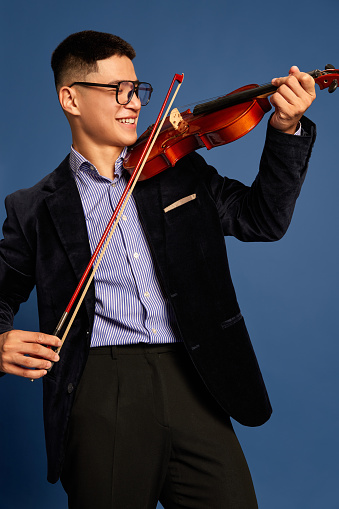 Portrait of aged 16-17 years old with brown hair caucasian male violinist standing in front of white background wearing pants who is happy and holding violin