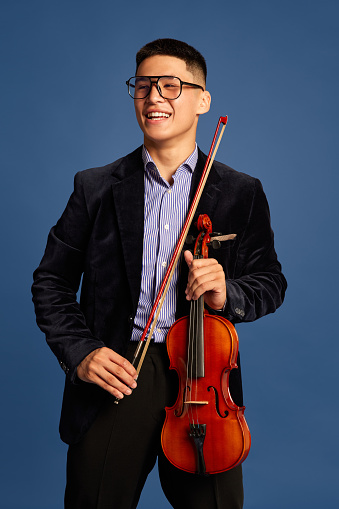 Portrait with handsome, young musicians, violinist wearing elegant suit and eyeglasses holding musical instrument and smiling over blue background. Concept of music, art, hobby, human emotions, ad