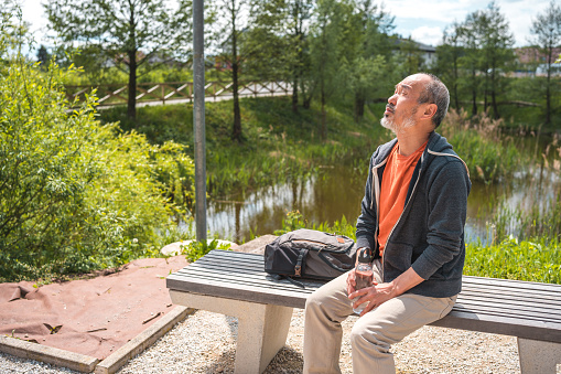 Mature Asian male sitting on a bench in a public park next to a pond. He is using a reusable water bottle.