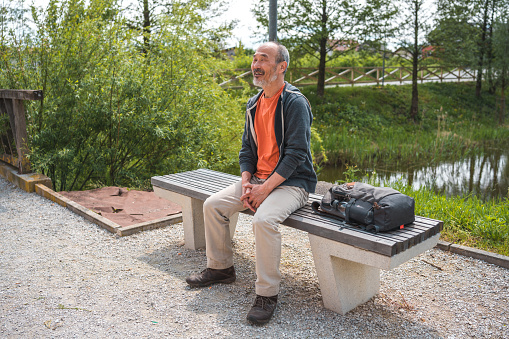 Mature Asian male sitting on a bench in a public park next to a pond. Spending some alone time outdoors enjoying the sun.