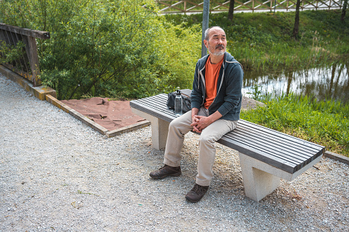 Mature Asian male sitting on a bench in a public park next to a pond. He is using a reusable water bottle.