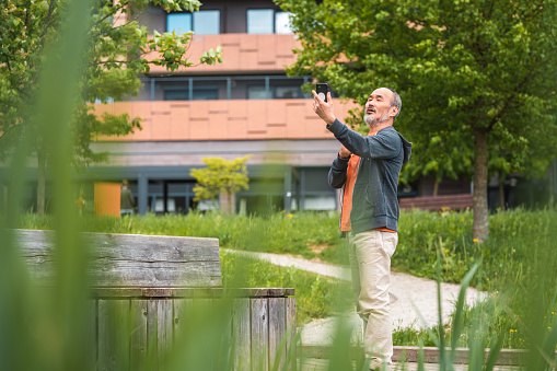 Cheerful mature Asian male standing in a public park. He is taking a selfie using a smartphone.
