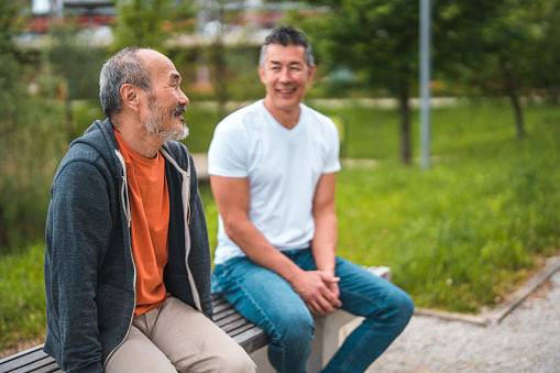 Cheerful mature Asian friends sitting on a bench in a public park. Having a fun spending time together outdoors.