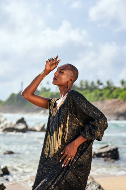 istock Epatage lgbtq black male posing with hands on camera on scenic ocean beach. Non-binary ethnic fashion model in long posh dress wears jewellery stands gracefully on sea shore and lighthouse. 1489460924