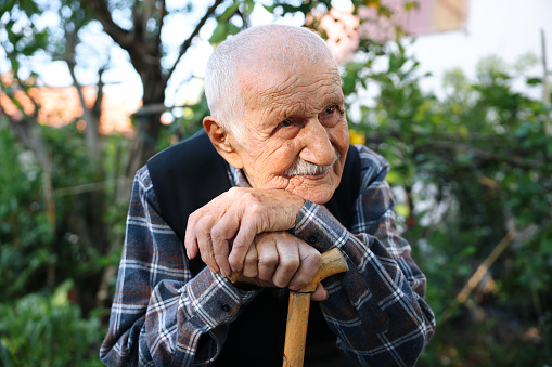 Old senior man with hands over the cane sitting in public park with sad expression. Elderly retiree with back pain using walking stick. Tree and flowers on background