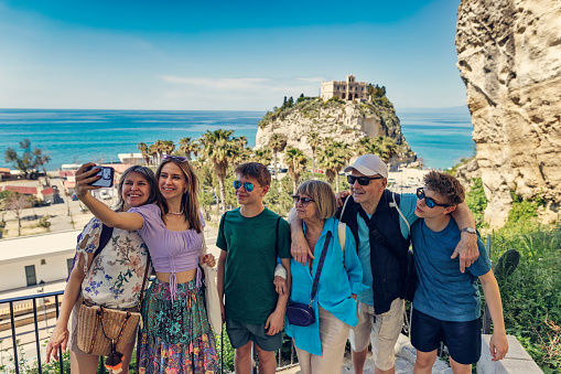 Teenagers with mother and grandparents are enjoying sunny spring day in Calabrian town of Tropea. They are taking selfies on the view point. Behind them there is a stunning cliff with Santuario di Santa Maria dell'Isola di Tropea.
Springtime, off-season vacations day in Tropea, Catania, Italy.
Shot with Canon R5