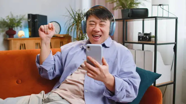 Photo of Happy asian man in wireless headphones relaxing sitting on sofa at home listening favorite music