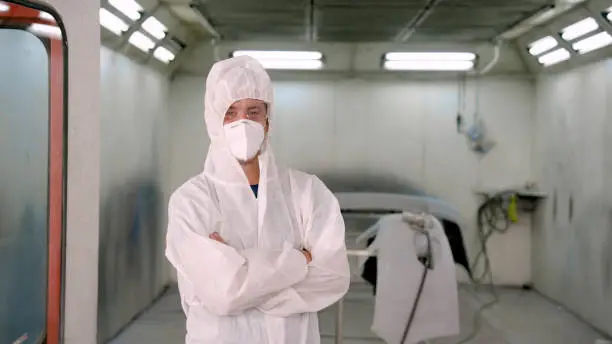 Caucasian males in white protective PPE suits standing five workshops of factory standard car paintwork. Wearing an anti-fog mask and goggles, I heard him cross his arms while repairing the car.