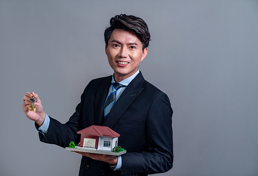 Confident Asian businessman holding house model, advertising home loan with smile. Real estate agent with sample house model in hand on isolated background for housing business advertisement. Jubilant