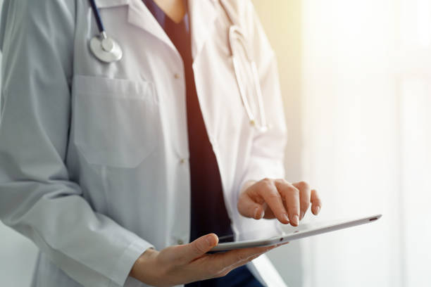 istock Doctor woman using tablet computer while standing near panorama window in clinic, close up. Physician or surgeon at work. Medicine concept 1489475576