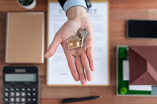 Top view key on real estate agent hand over wooden table surrounded by calculator and legal document as symbolic for house loan or mortgage, offering with persuasive gesture for home sales. Jubilant