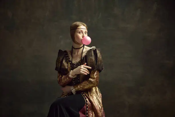 Portrait of pretty young girl, medieval princess in vintage dress posing with bubble gum against dark green background. Concept of history, renaissance art remake, comparison of eras, modernity