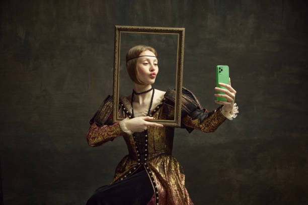 portrait of pretty young girl, royal person in vintage dress holding picture frame and taking selfie with phone against dark green background. - princess women duchesses renaissance imagens e fotografias de stock