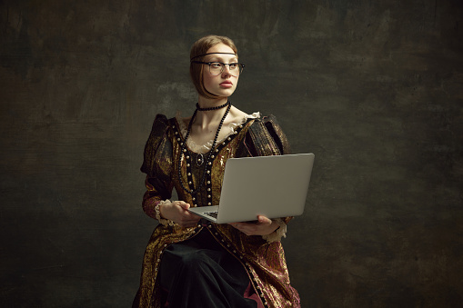 Portrait of young pretty girl, queen in vintage dress posing with laptop over dark green background. Education, freelance job, business. Concept of history, renaissance art remake, comparison of eras