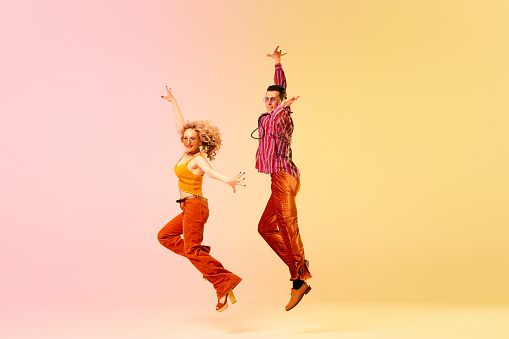 Dynamic image of beautiful young couple, man and woman in stylish vintage clothes dancing against gradient pink yellow background. Concept of retro style, dance, fashion, art, hobby, music, 70s