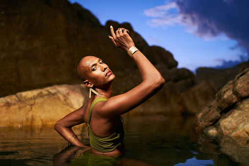 Androgynous bipoc lgbtq model poses in water inside natural pool at night. Non-binary person shows jewelry - rings with gems on fingers, brass nose ring, golden earrings, bracelets, stands in a pond.