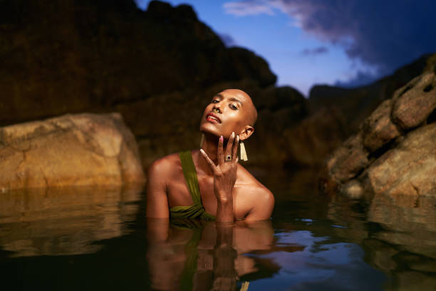 istock Androgynous bipoc lgbtq model poses in water inside natural pool at night. Non-binary person shows jewelry - rings with gems on fingers, brass nose ring, golden earrings, bracelets, stands in pond. 1489486129