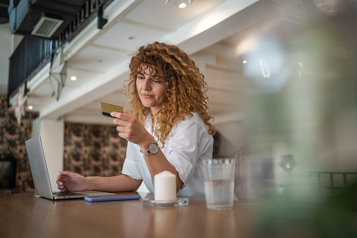 A young woman is sitting in a coffee shop and while drinking coffee she is shopping online, ordering products on her laptop and paying with a payment card online