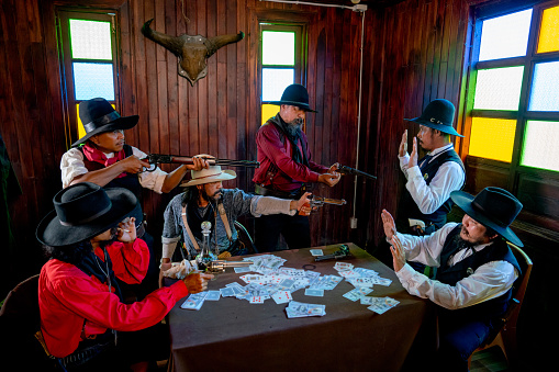 Group of cowboy men hold gun and point to each other during play card in room.