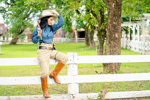 Wide shot of pretty Asian woman with cowboy style hold rifle gun on her shoulder and sit on fence in farm with day light.