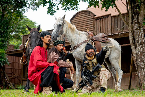 Group of men with soldier and arab clothes sit down in front of horse and old building look like discuss plan to fighting with enemy.