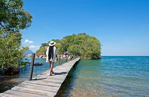 Woman strolls on elevated walkway jetty between Mumbo and Jumbo islands, Mumbo Island, Lake Malawi, Malawi, Africa. Mumbo Island, within Lake Malawi National Park, is in the Salima District 100 kilometers east of the capital Lilongwe. Malawi, the landlocked country in southeastern Africa, is a country of highlands split by the Great Rift Valley and the huge Lake Malawi, whose southern end is within Lake Malawi National Park and several other parks are now habitat for diverse wildlife from colorful fish to the Big Five. Cape Maclear is known for its beach resorts, whilst several islands offer rest and recreation.