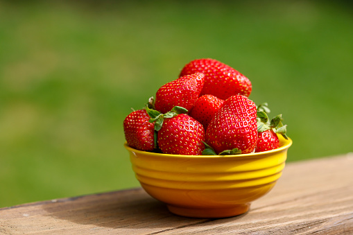 A bowl of fresh red strawberries in a yellow bowl sitting on a deck with a green grass background