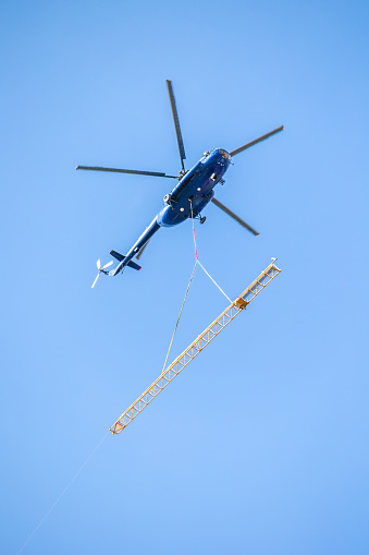 Dark blue helicopter seen from bellow with yellow segment of construction crane hanging on rope, flying against clear blue sky