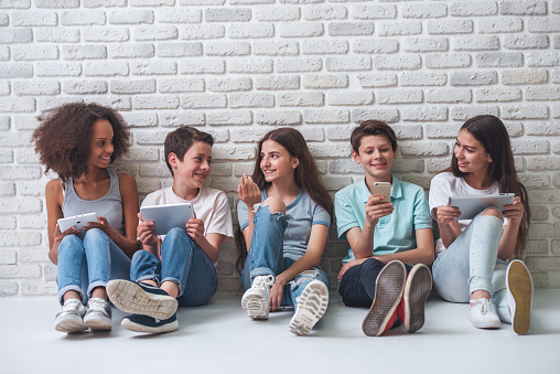 Group of teenage boys and girls is using gadgets, talking and smiling, sitting against white brick wall