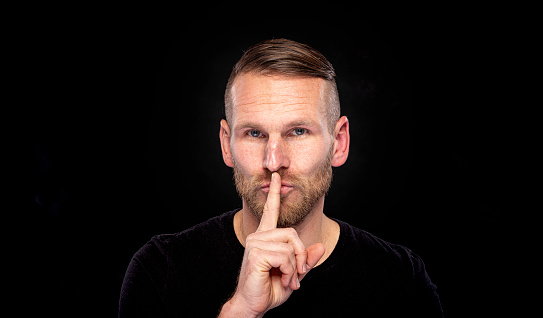 A man holds a finger to his mouth showing a sign of silence, on a dark background.